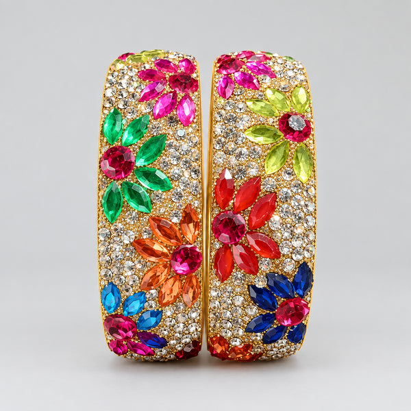 Two ornate bangles with colorful gemstones held by a white gloved hand against a wooden background.